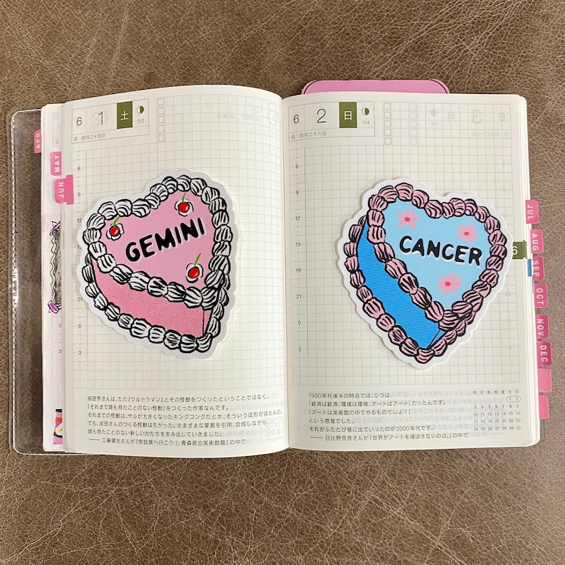 May include: Two heart-shaped cake stickers with pink and blue frosting. The pink cake has the word "GEMINI" on it and the blue cake has the word "CANCER" on it.