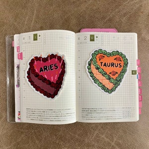 May include: Two heart-shaped birthday cake stickers with the zodiac signs Aries and Taurus. The Aries cake is red with pink frosting and candles. The Taurus cake is orange with green frosting and red roses.