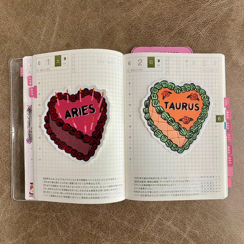 May include: Two heart-shaped birthday cake stickers with the zodiac signs Aries and Taurus. The Aries cake is red with pink frosting and candles. The Taurus cake is orange with green frosting and red roses.