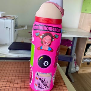 May include: Pink reusable water bottle with a variety of stickers, including a sticker that says "femininoomenon!" and a sticker that says "Hot Girls Read Books".