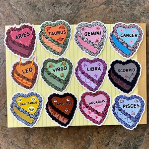 May include: Twelve heart-shaped stickers featuring the astrological signs. Each sticker is a different color and has a different design. The signs are: Aries, Taurus, Gemini, Cancer, Leo, Virgo, Libra, Scorpio, Sagittarius, Capricorn, Aquarius, and Pisces.
