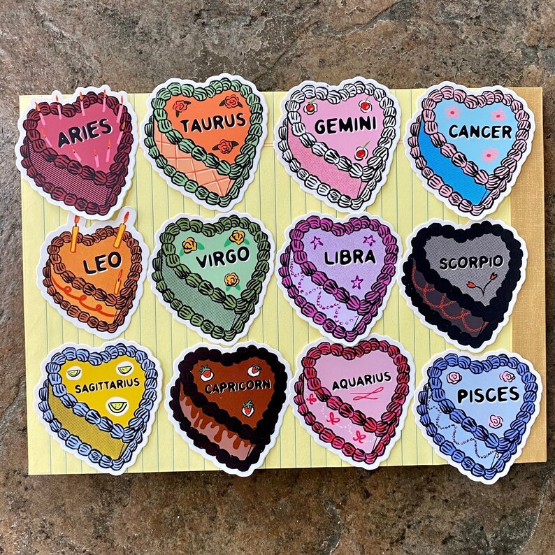 May include: Twelve heart-shaped stickers featuring the astrological signs. Each sticker is a different color and has a different design. The signs are: Aries, Taurus, Gemini, Cancer, Leo, Virgo, Libra, Scorpio, Sagittarius, Capricorn, Aquarius, and Pisces.