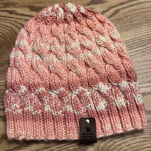 May include: A handmade cable-knit beanie in shades of pink and cream. The hat features a textured pattern and a ribbed brim. A small leather tag with the text "Handmade knits by Victoria" is attached.