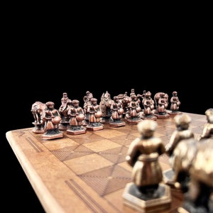 May include: A close-up of a chessboard with copper-colored chess pieces. The pieces are arranged in a line on the board, with the king and queen in the center. The board is made of wood and has a dark brown finish.