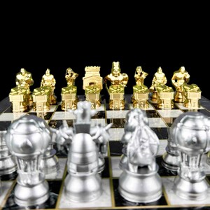 May include: A gold chess set with silver chess pieces on a black and white chessboard. The gold pieces are in the starting position, while the silver pieces are in the middle of the board.