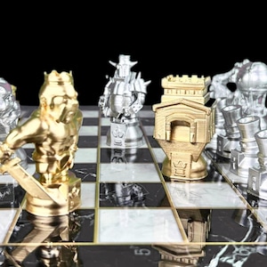 May include: A gold and silver chess set with a black and white marble board. The pieces are stylised and include a king, queen, knight, and rook. The board is set up for a game of chess.