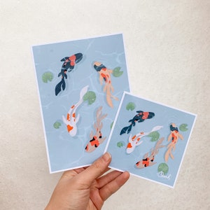 May include: Two prints of colorful koi fish swimming in a blue pond with lily pads. The larger print has four fish and the smaller print has three fish. Both prints have a light blue background and are signed "Oanh" in the bottom right corner.