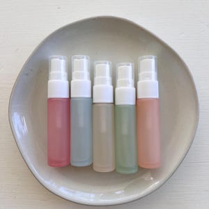 Könnte beinhalten: Fünf kleine, gefrostete Glassprühflaschen mit weißen Verschlüssen. Die Flaschen sind in einem Kreis auf einem weißen Teller angeordnet. Die Flaschen sind in den folgenden Farben: rosa, blau, beige, grün und pfirsich.