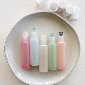 May include: Five frosted glass roller bottles with different colored liquids and metal roller balls. The bottles are arranged in a row on a white ceramic plate.