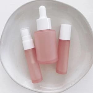 Puede incluir: Tres botellas de vidrio con tapas blancas y líquido rosa. Las botellas están en un plato blanco.