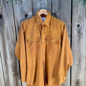 May include: A long-sleeved, button-down shirt in a warm, golden-brown hue. The shirt features two chest pockets with button closures, a classic collar, and buttoned cuffs. The shirt is hanging on a wooden hanger.