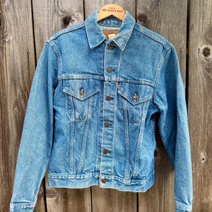 May include: A light blue denim jacket with a classic design, featuring a button-up front, two chest pockets, and a collar. The jacket is hanging on a wooden hanger against a weathered wooden background. The Levi's brand red tag is visible.