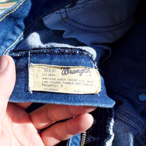 May include: Close-up of a blue denim Wrangler jeans label with white text. The label reads "30X30 410 SNVR MACHINE WASH INSIDE OUT WITH LIKE COLORS, TUMBLE DRY, REMOVE PROMPTLY R 5F0489809 MADE IN U.S.A."