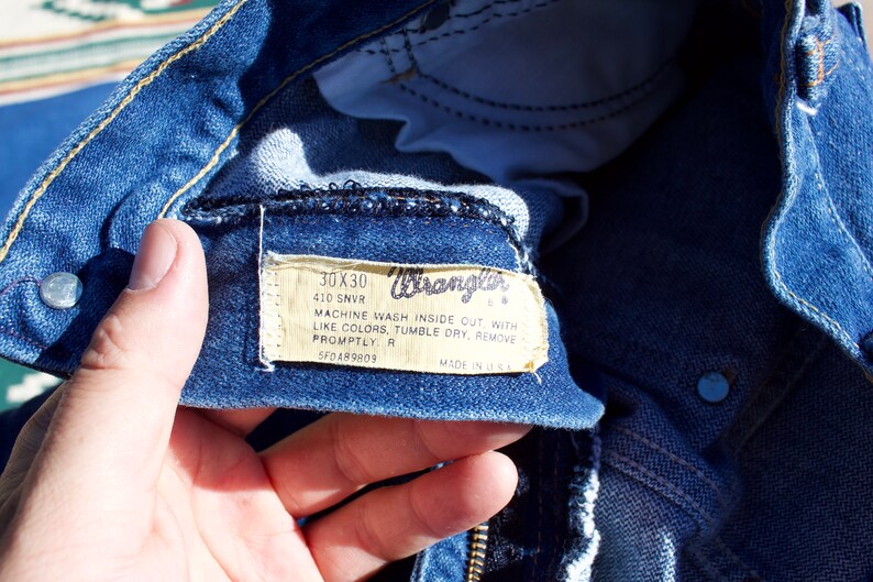 May include: Close-up of a blue denim Wrangler jeans label with white text. The label reads "30X30 410 SNVR MACHINE WASH INSIDE OUT WITH LIKE COLORS, TUMBLE DRY, REMOVE PROMPTLY R 5F0489809 MADE IN U.S.A."