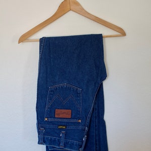 May include: A pair of blue denim jeans folded and hanging on a wooden hanger. The jeans have a brown leather patch with the text "Wrangler" on the back pocket.