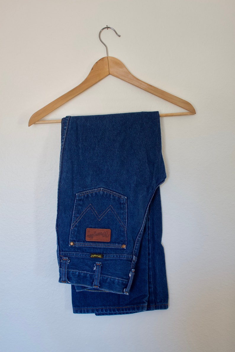 May include: A pair of blue denim jeans folded and hanging on a wooden hanger. The jeans have a brown leather patch with the text "Wrangler" on the back pocket.