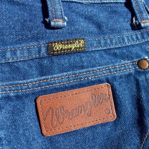 May include: A pair of blue denim jeans with a brown leather patch that says "Wrangler" on it. The jeans have a small gold button on the back pocket.