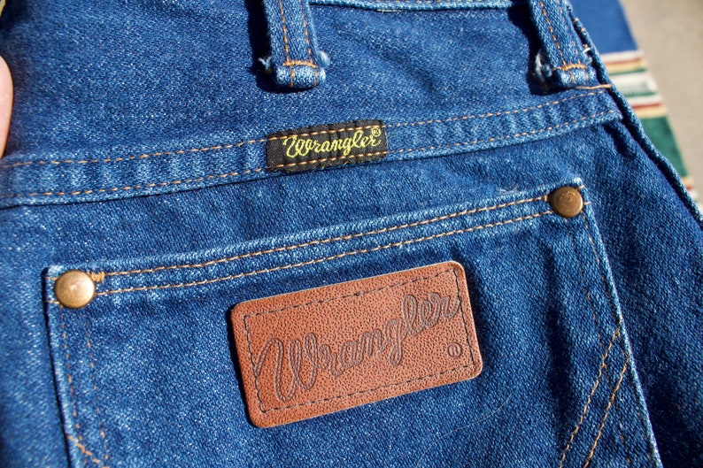 May include: A pair of blue denim jeans with a brown leather patch that says "Wrangler" on it. The jeans have a small gold button on the back pocket.