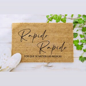 May include: A rectangular coir doormat in tan. The mat has the words "Rapido Rapido" in black script, with more text underneath. White trainers are visible on the left. Green ivy is on the right.
