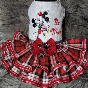 Pu&ograve; includere: Un abito bianco in pizzo per cani con una gonna a quadri rossi e neri. L'abito presenta un'immagine ricamata di Topolino e Minnie Mouse che si baciano con il testo "Be Mine".