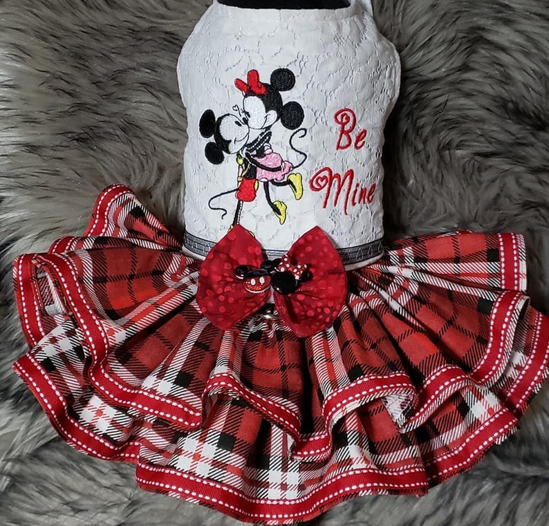 Pu&ograve; includere: Un abito bianco in pizzo per cani con una gonna a quadri rossi e neri. L'abito presenta un'immagine ricamata di Topolino e Minnie Mouse che si baciano con il testo "Be Mine".