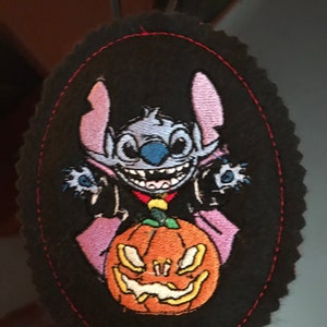 May include: A black felt oval ornament with a stitched border. The ornament features an embroidered image of Stitch from Lilo & Stitch dressed as a vampire holding a jack-o'-lantern.
