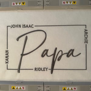 May include: A white fabric piece secured in an embroidery hoop featuring the word Papa in large grey cursive script. A thin grey rectangular border surrounds the text, with the names John Isaac, Kanan, Ridley, and Archie embroidered along the sides.