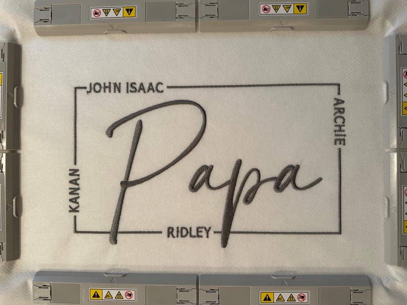 May include: A white fabric piece secured in an embroidery hoop featuring the word Papa in large grey cursive script. A thin grey rectangular border surrounds the text, with the names John Isaac, Kanan, Ridley, and Archie embroidered along the sides.