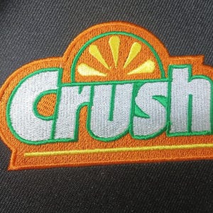May include: A circular black fabric patch featuring an embroidered logo with the word Crush in silver letters. The logo is outlined in green and orange, with a stylized yellow orange slice graphic positioned above the text on a black background.