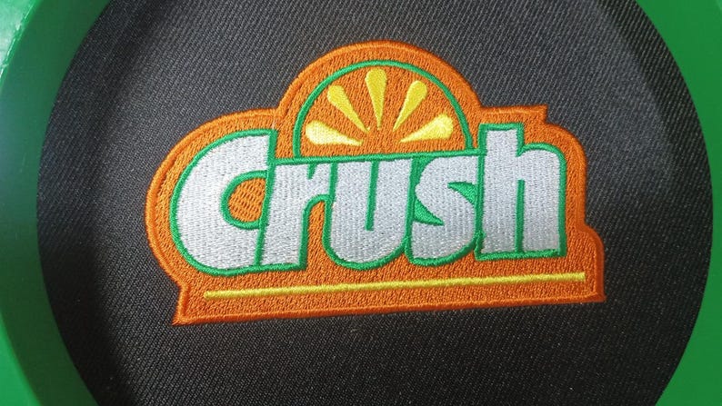 May include: A circular black fabric patch featuring an embroidered logo with the word Crush in silver letters. The logo is outlined in green and orange, with a stylized yellow orange slice graphic positioned above the text on a black background.