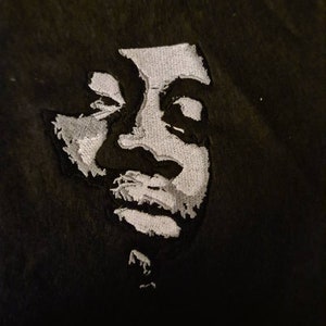 May include: A black fabric patch featuring a high-contrast, white embroidered portrait of a person. The embroidery uses dense stitching to create light and shadow effects, forming the facial features, hair, and chin against the dark background.