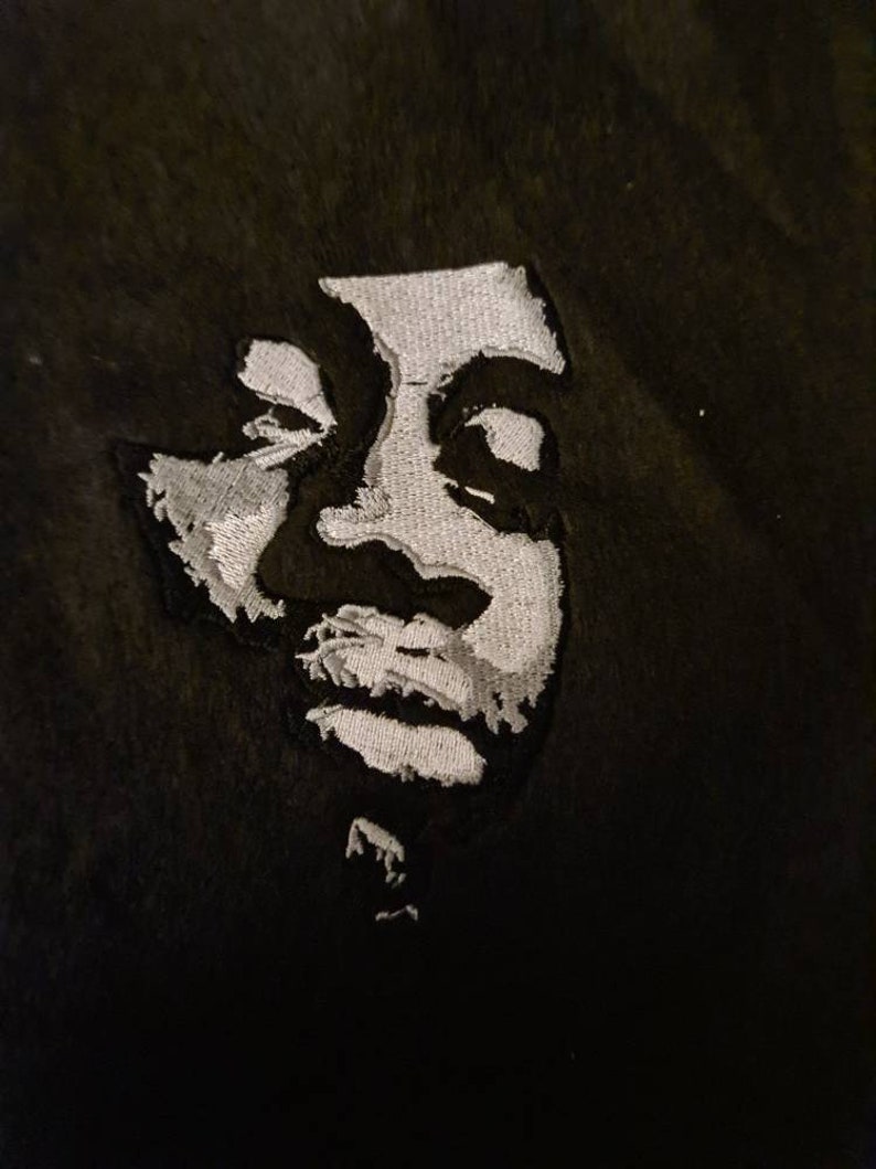 May include: A black fabric patch featuring a high-contrast, white embroidered portrait of a person. The embroidery uses dense stitching to create light and shadow effects, forming the facial features, hair, and chin against the dark background.