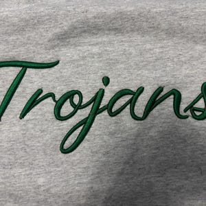 May include: A light gray heathered fabric garment featuring the word Trojans embroidered in a dark green cursive script font. The embroidery has a raised texture and a satin-like finish, standing out clearly against the soft, textured gray background.