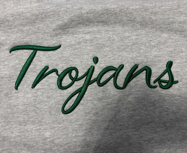 May include: A light gray heathered fabric garment featuring the word Trojans embroidered in a dark green cursive script font. The embroidery has a raised texture and a satin-like finish, standing out clearly against the soft, textured gray background.