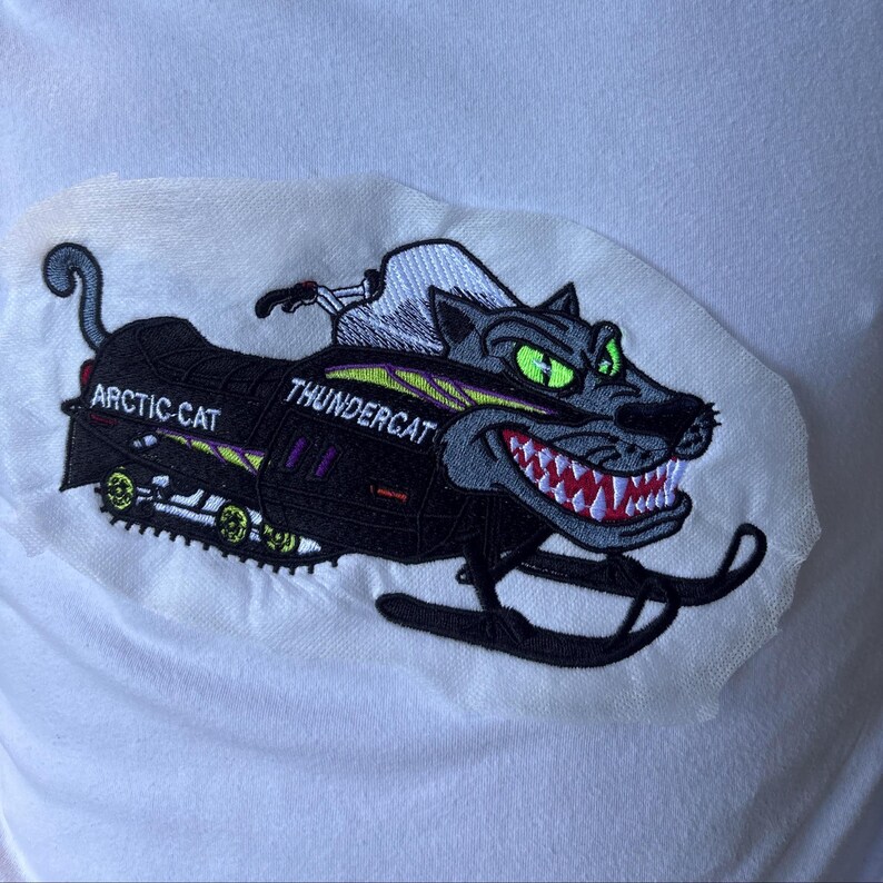 May include: An embroidered patch on a white fabric background featuring a black snowmobile with a stylized cat head design. The text ARCTIC CAT and THUNDERCAT is visible. The cat has bright green eyes, sharp white teeth, and red gums.