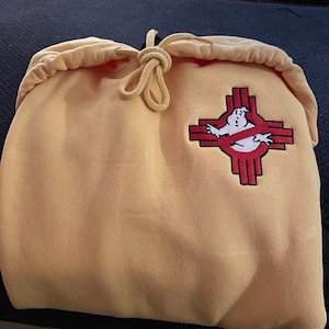 May include: A light orange hooded sweatshirt featuring a drawstring hood and an embroidered patch on the chest. The patch displays a white ghost character inside a red, cross-shaped symbol, combining a classic ghost icon with a geometric design.