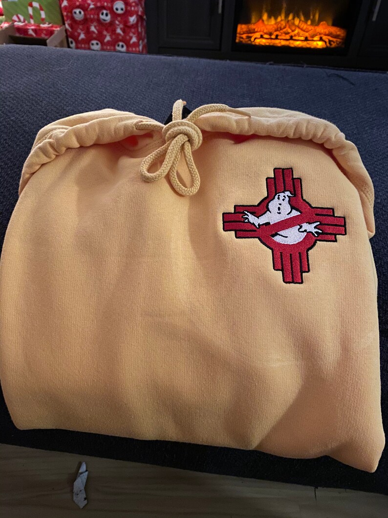 May include: A light orange hooded sweatshirt featuring a drawstring hood and an embroidered patch on the chest. The patch displays a white ghost character inside a red, cross-shaped symbol, combining a classic ghost icon with a geometric design.