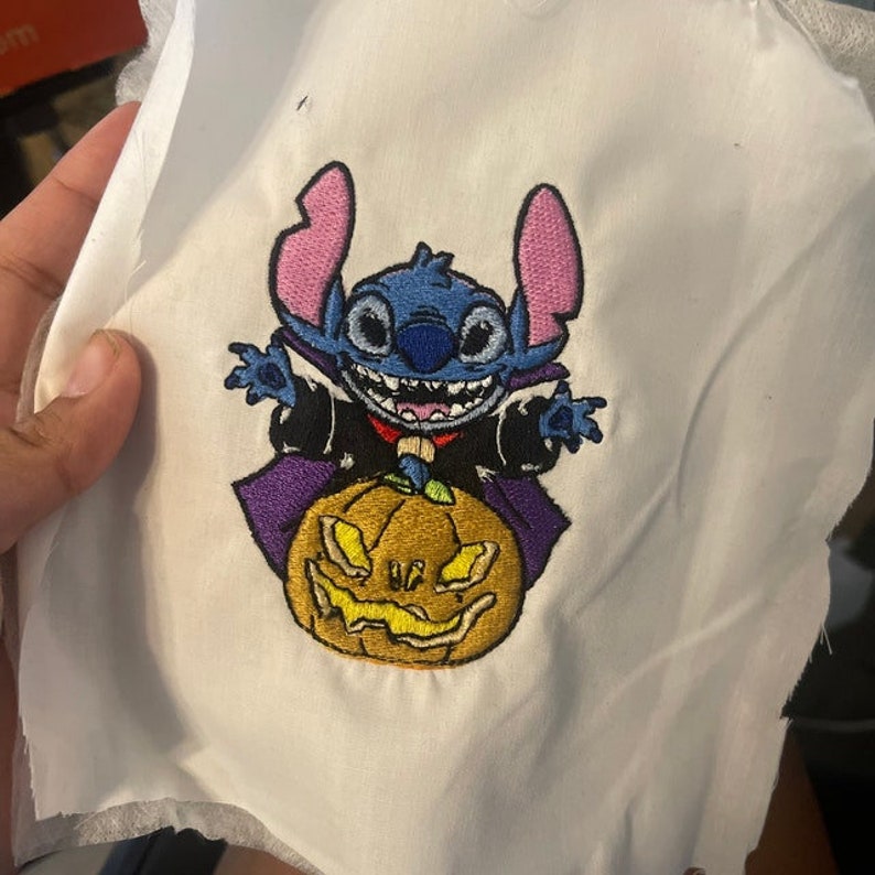 May include: An embroidered design featuring Stitch, a cartoon character, dressed as a vampire, sitting on top of a jack-o'-lantern.