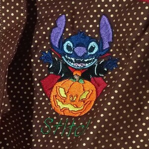 May include: A brown fabric with gold polka dots features an embroidered image of Stitch, a blue alien character, dressed as a vampire and holding a jack-o'-lantern. The text "Stitch!" is embroidered below the image.