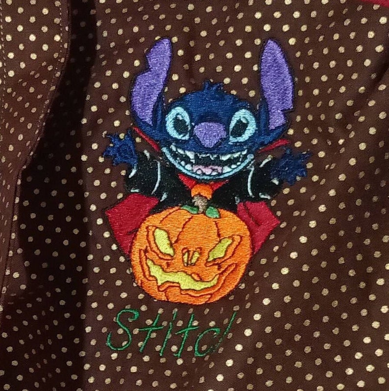 May include: A brown fabric with gold polka dots features an embroidered image of Stitch, a blue alien character, dressed as a vampire and holding a jack-o'-lantern. The text "Stitch!" is embroidered below the image.