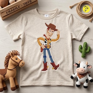 Może przedstawiać: Beżowy t-shirt z kr&oacute;tkim rękawem z wyhaftowaną grafiką kowboja w brązowym kapeluszu, ż&oacute;łtej koszuli w kratę, kamizelce w łaty, niebieskich spodniach i brązowych butach. Koszulka ma okrągły dekolt oraz metkę z rozmiarem 4T przy kołnierzu.