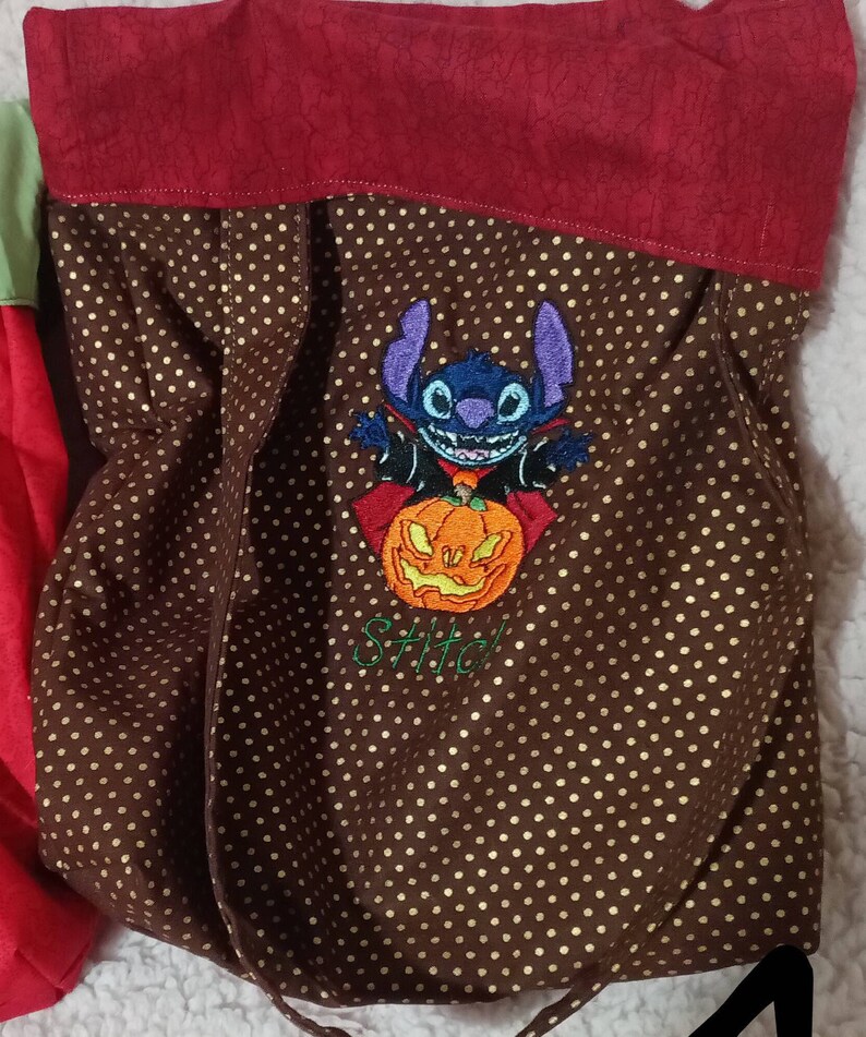May include: A brown fabric trick-or-treat bag with gold polka dots. The bag has a red fabric top and an embroidered image of Stitch from Lilo & Stitch dressed as a vampire holding a jack-o-lantern. The text "Stitch!" is embroidered below the image.
