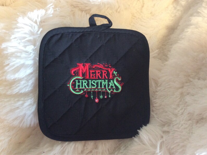 Pu&ograve; includere: Presa pentola imbottita nera con un ricamo rosso e verde "Buon Natale" con ornamenti appesi.