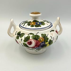 May include: A white ceramic sugar bowl with a lid, decorated with hand-painted floral patterns in red, pink, yellow, blue, and green. The bowl has two handles and a gold-coloured knob on the lid. The floral pattern is repeated on the lid.