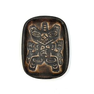 Peut inclure: Un plat en céramique ovale brun foncé avec un motif amérindien sculpté. Le motif présente une figure centrale avec un visage stylisé et deux figures latérales. Le plat a une surface texturée et est posé sur un fond blanc.