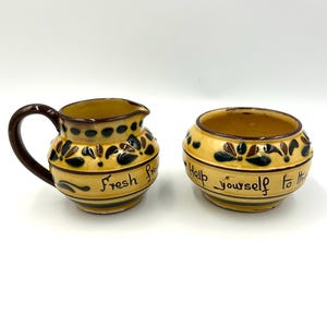 May include: A yellow ceramic creamer and sugar bowl set. The creamer has a brown handle and the words "Fresh Cream" are written on the side. The sugar bowl has the words "Help yourself to this" written on the side. Both pieces have a floral design.