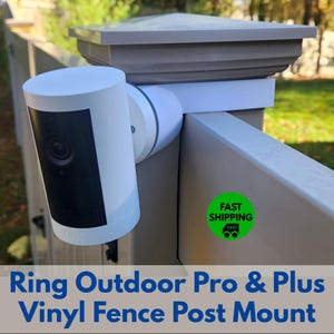May include: A white and black security camera mounted on a vinyl fence post. The camera is cylindrical with a black front panel. The text "Ring Outdoor Pro & Plus Vinyl Fence Post Mount" is displayed below the camera.