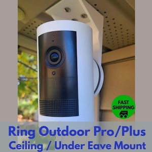 May include: White and black security camera mounted on a white bracket. The camera has a black lens and a speaker grill at the bottom. The text "Ring Outdoor Pro/Plus Ceiling / Under Eave Mount" is at the bottom.