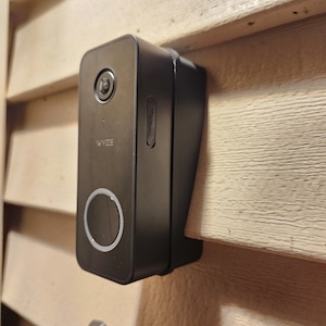 May include: A black Wyze video doorbell with a round button and a micro SD card slot. The doorbell is mounted on a light brown wooden surface.
