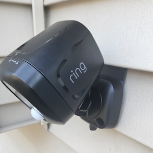 May include: A black Ring security camera with a motion sensor and a white light. The camera is mounted on a white siding.